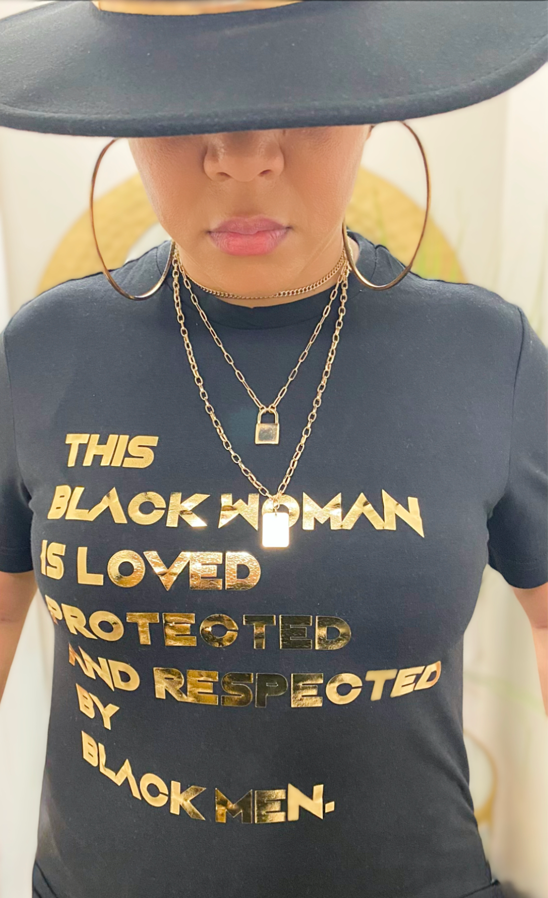 Black Tshirt. Gold foil letters that read "THIS BLACK WOMAN IS LOVED PROTECTED AND RESPECTED BY BLACK MEN" in all caps. Worn by a woman with gold necklaces, gold hoops, and a black hat.